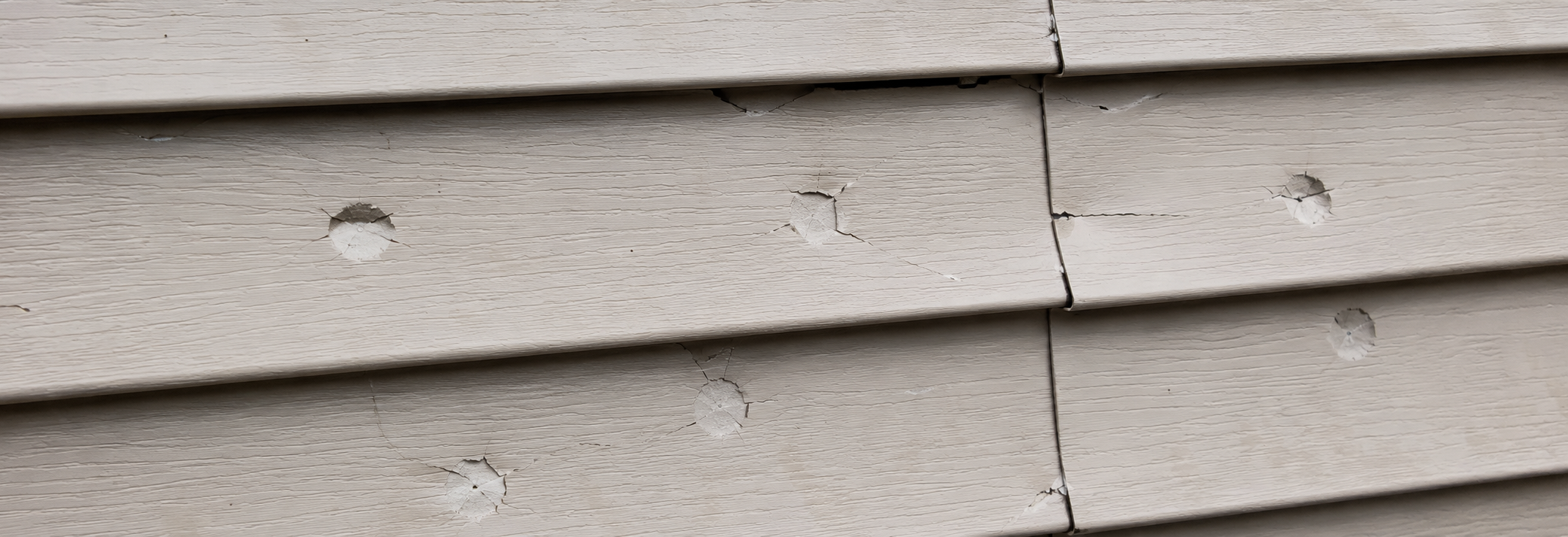 Close-up of vinyl siding showing circular hail impact cracks and cracked seam joints in raking sunlight
